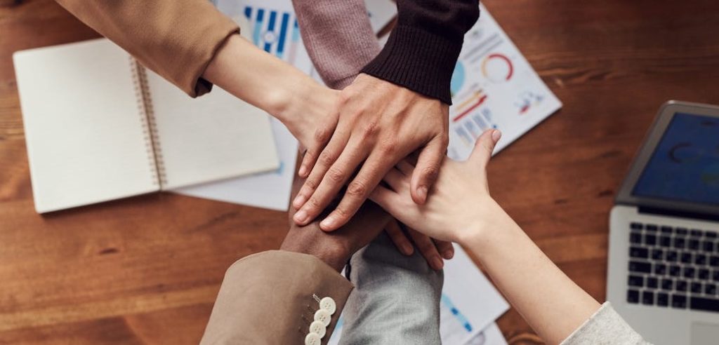 Raison d’être d’une entreprise : Gros plan sur plusieurs mains empilées au centre d'une table, symbolisant la cohésion et le travail d’équipe, avec en arrière-plan un ordinateur portable, un carnet ouvert et des documents de travail comportant des graphiques colorés.